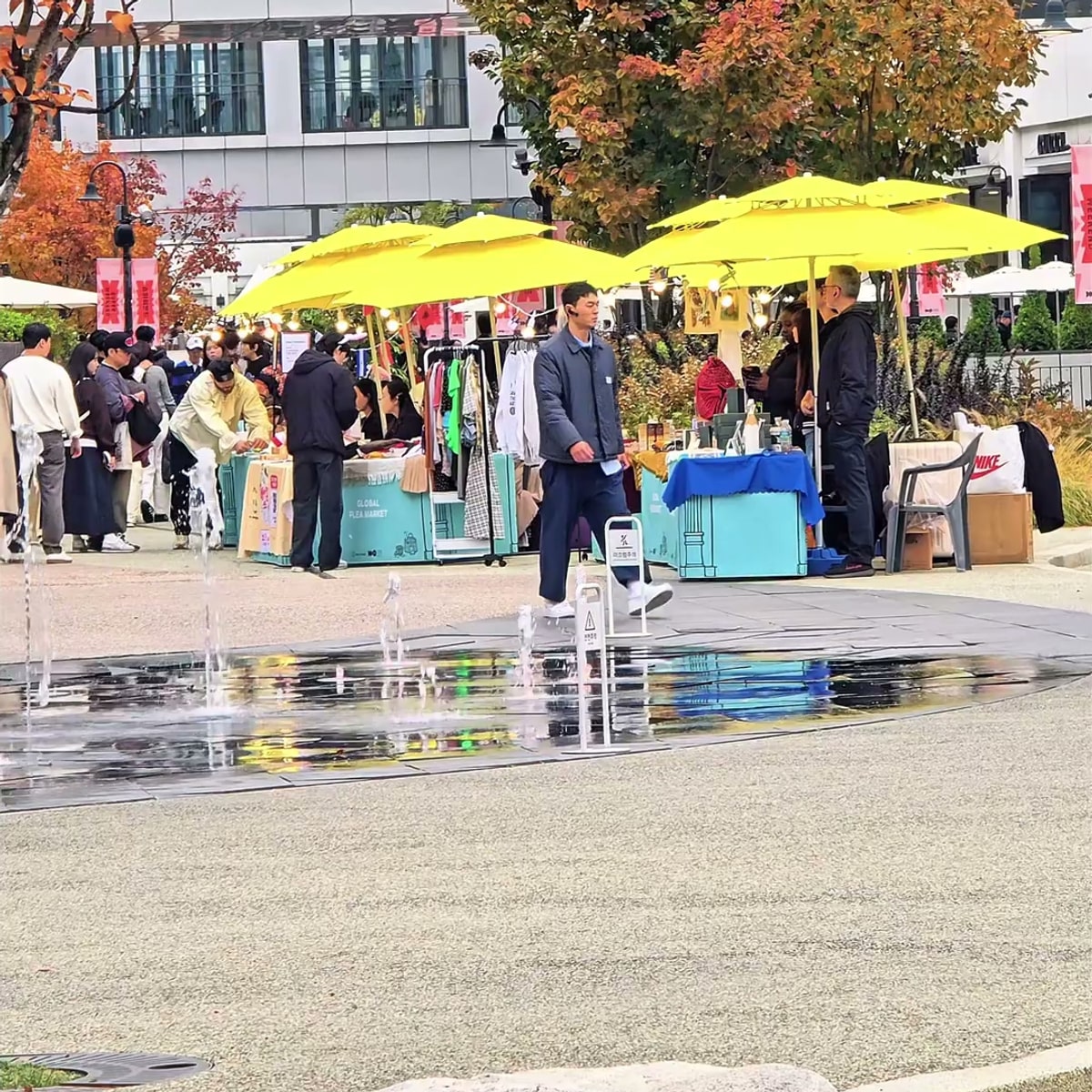 야외 광장에 노란 파라솔을 얹은 민트색 부스가 줄지어 서 있고 방문객들이 매대를 둘러보는 플리마켓 현장, 앞쪽에는 바닥 분수가 솟아 있는 장면