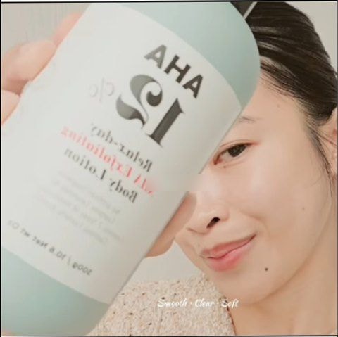 크리에이터가 얼굴 옆으로 민트색 AHA 12% 바디로션 용기를 들어 라벨이 보이게 촬영한 클로즈업 장면. 아래쪽에 “Smooth · Clear · Soft”라는 자막이 있다