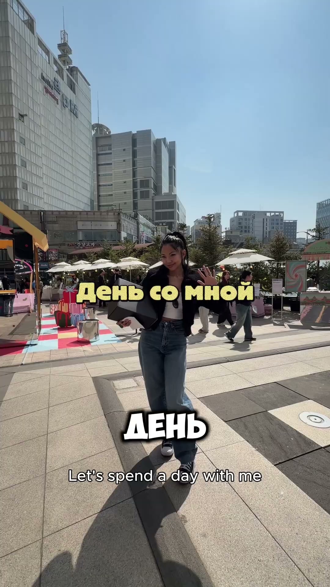 행사장 광장으로 걸어 들어오며 손을 흔드는 크리에이터의 세로 영상 장면. 화면에 러시아어 자막과 영어 자막이 함께 붙어 있고 뒤로 파라솔과 마켓 부스가 늘어서 있다