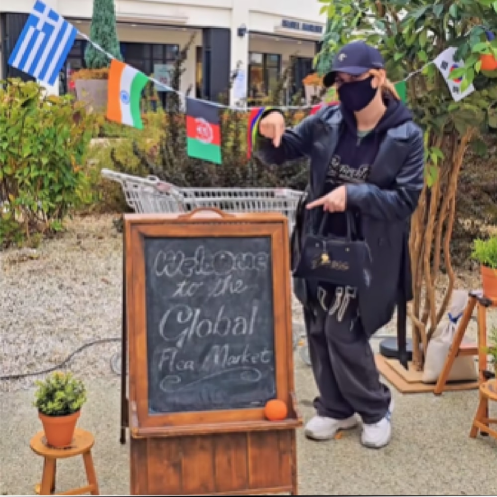 여러 나라 국기가 걸린 야외 행사장 입구에서 크리에이터가 ‘Welcome to the Global Flea Market’이라고 분필로 쓰인 칠판 입간판을 손가락으로 가리키는 장면