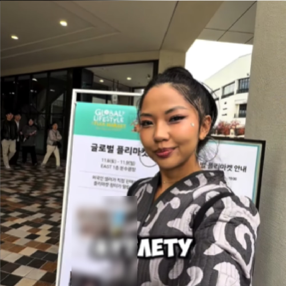 행사장 입구에 세워진 ‘글로벌 플리마켓’ 안내 배너 앞에서 크리에이터가 카메라를 보고 서 있는 장면. 화면 아래에 러시아어 자막이 얹혀 있다