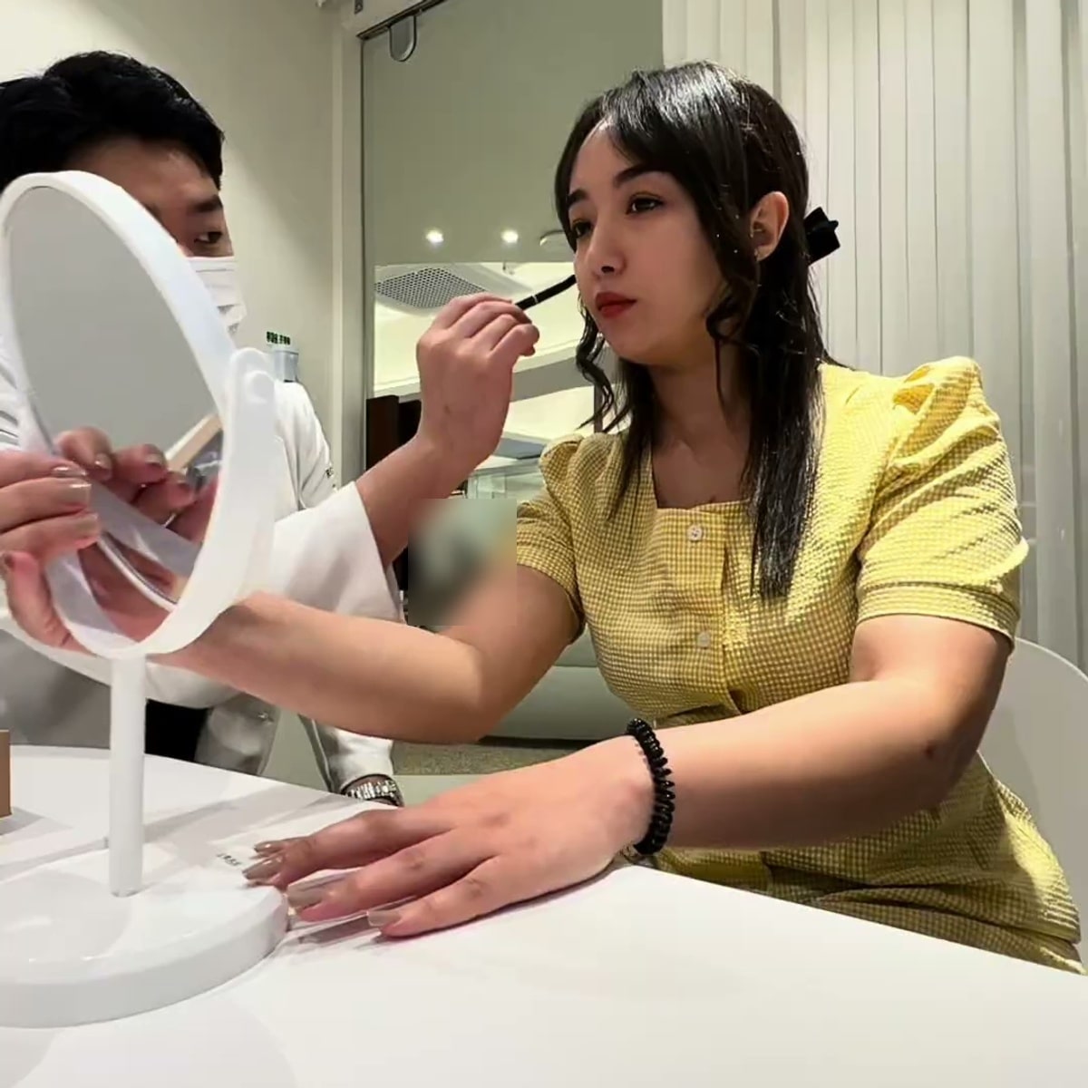 노란 원피스를 입은 크리에이터가 시술 베드 옆 테이블에 앉아 거울을 보고, 흰 가운을 입은 의료진이 거울을 들고 얼굴 부위를 펜슬로 짚어 설명하는 장면