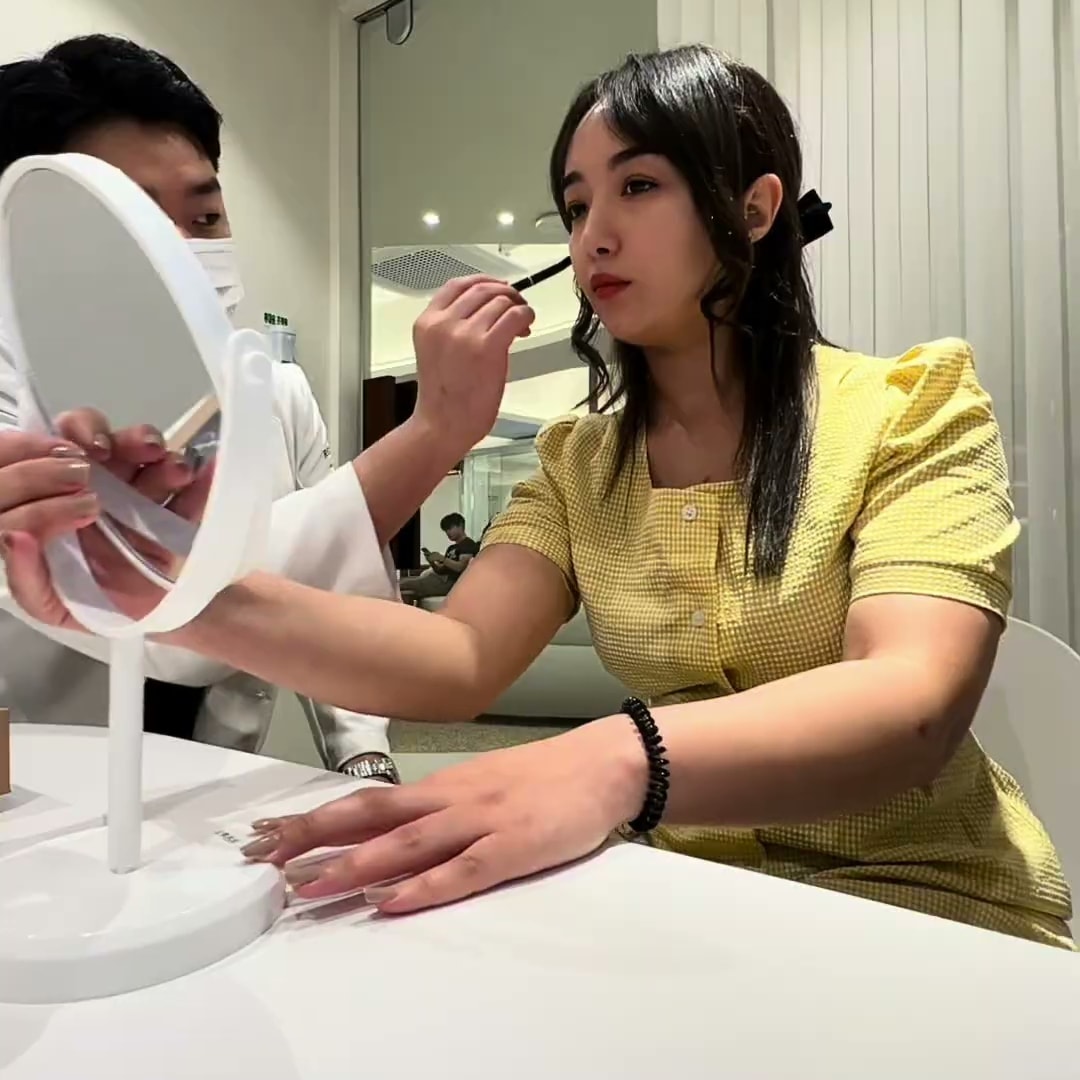 시술을 마친 크리에이터가 거울 앞에 앉아 담당자에게 눈썹 부위 마무리 손질을 받는 장면. 밝은 흰색 상담 공간이다