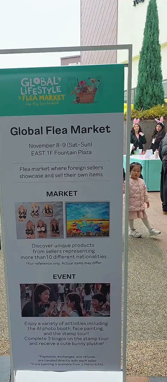 행사장 입구에 세워진 세로형 안내 입간판. ‘Global Flea Market, November 8–9 (Sat–Sun), EAST 1F Fountain Plaza’와 10개국 이상의 셀러가 참여한다는 안내, 스탬프 투어·페이스 페인팅 등 이벤트 설명이 영어로 적혀 있다
