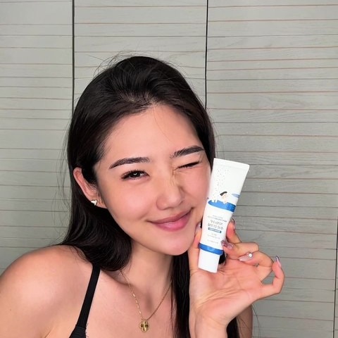 튜브형 화장품을 얼굴 옆에 들고 한쪽 눈을 찡긋하며 웃는 영어권 크리에이터의 실내 촬영 컷