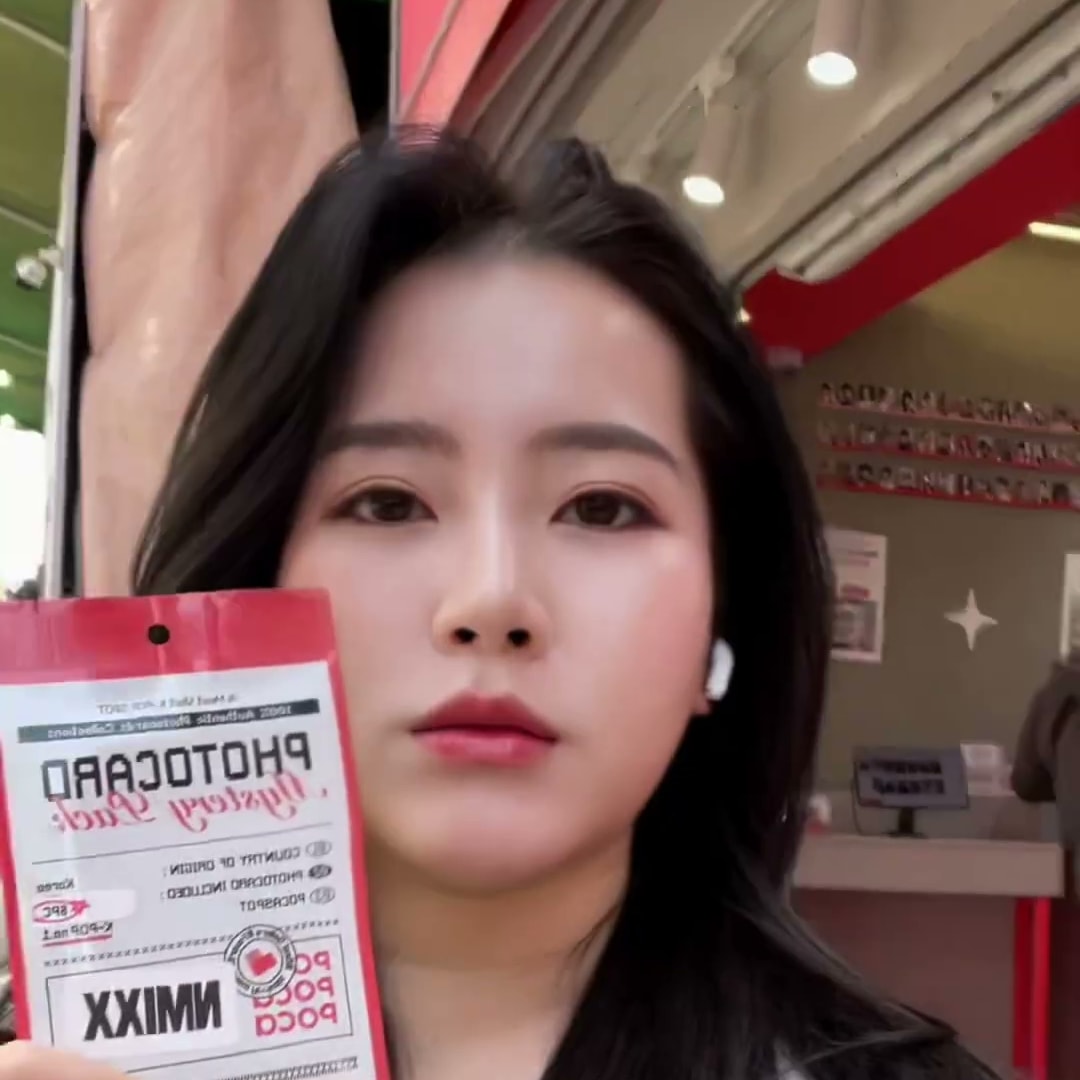 붉은 조명의 케이팝 굿즈 매장 안에서 포토카드 팩을 얼굴 옆에 들어 보이는 베트남 출신 크리에이터의 컷
