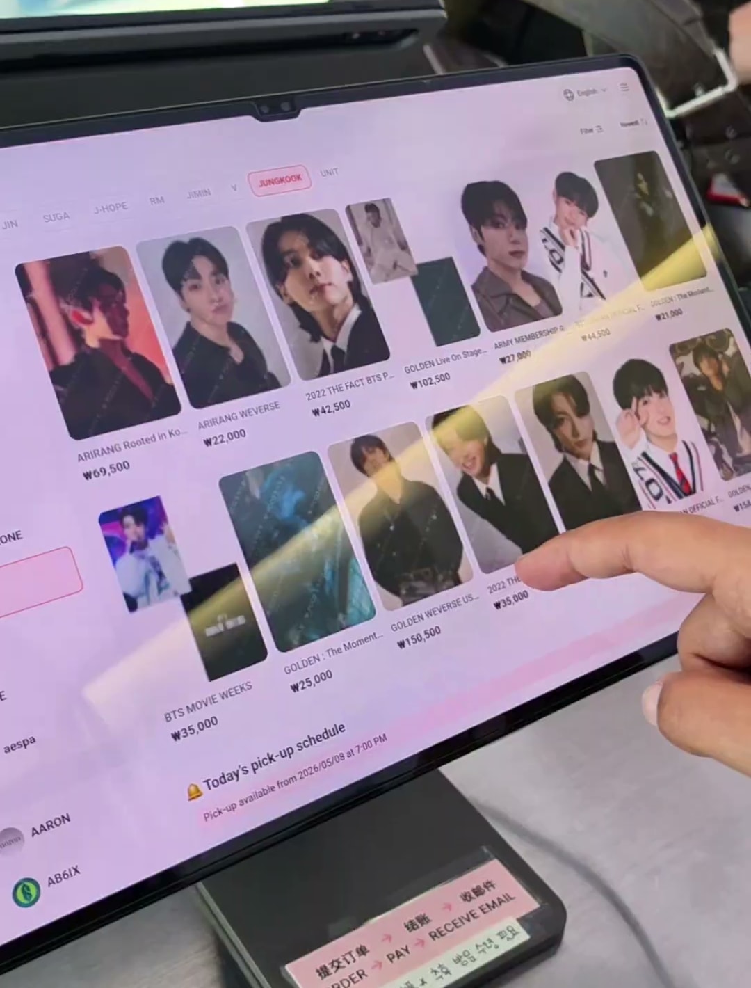 태블릿 주문 화면을 손가락으로 가리키는 장면. 오른쪽 위에 영어 언어 전환 메뉴가 있고 아래 안내판에는 주문·결제·수령 순서가 중국어와 영어와 한국어로 적혀 있다
