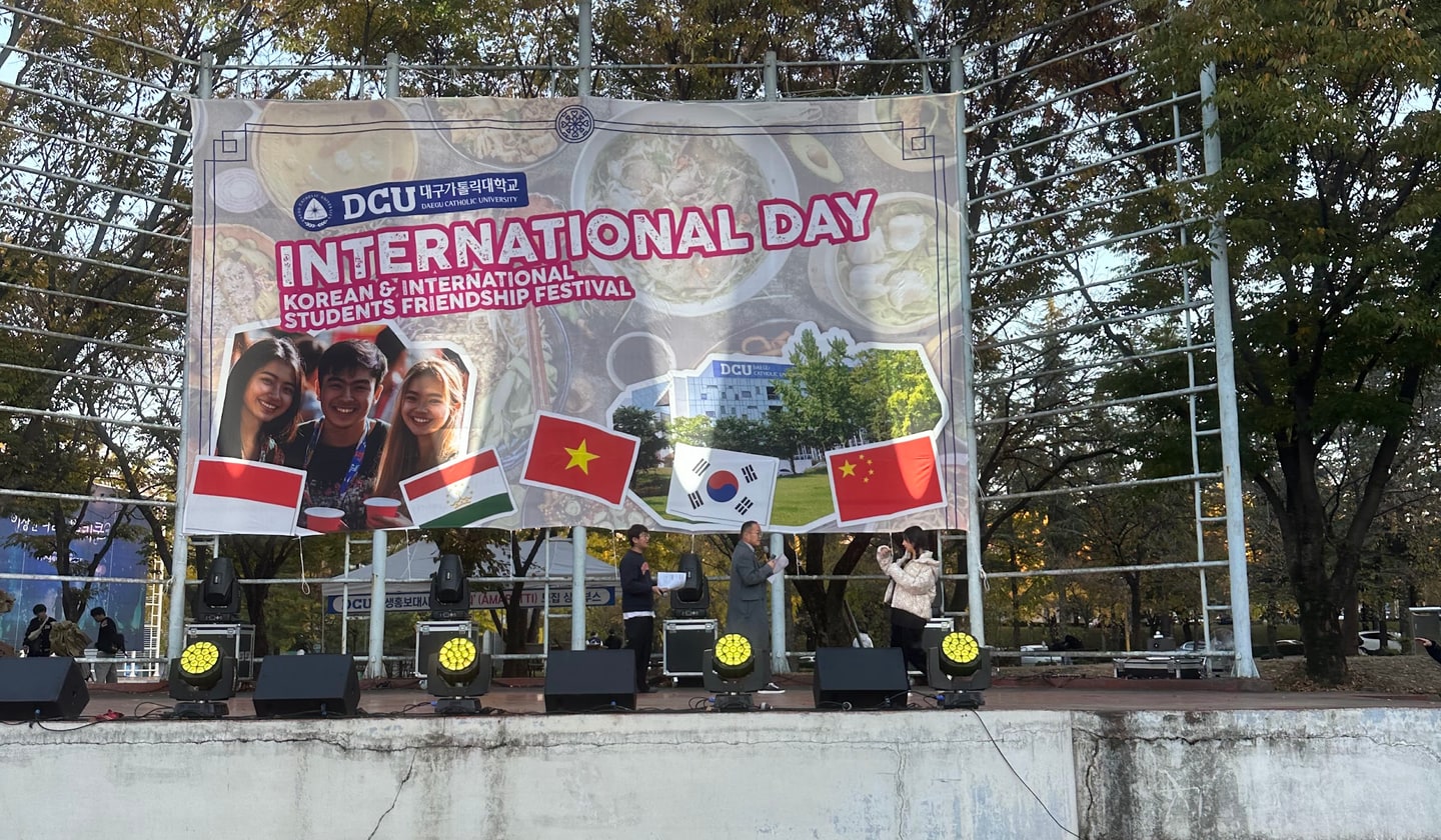 야외 무대에 걸린 대형 현수막에 대구가톨릭대학교 로고와 INTERNATIONAL DAY, KOREAN & INTERNATIONAL STUDENTS FRIENDSHIP FESTIVAL 문구가 적혀 있고 대한민국·베트남·중국·인도네시아 등 여러 나라 국기가 나란히 붙어 있다