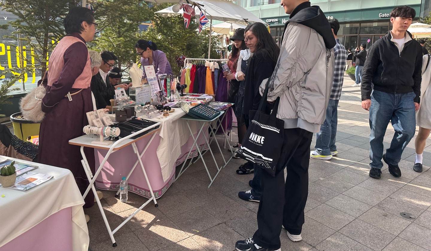 현대아울렛 동대문점 야외 광장에 분홍색 천을 두른 플리마켓 부스가 늘어서 있고, 한복 차림의 판매자가 액세서리 매대를 지키는 옆으로 여러 국적의 방문객이 줄지어 구경하고 있다