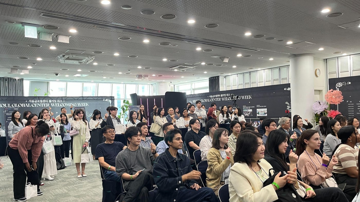 서울글로벌센터 웰커밍 위크 행사장에 다국적 참가자들이 모여 앉아 있고, 뒤쪽에도 사람들이 서서 지켜보고 있다.