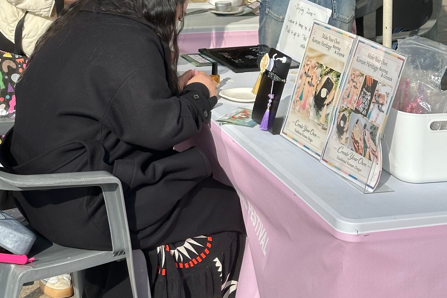야외 행사 체험 부스 테이블. 참가자가 등을 보인 채 앉아 키링을 만들고 있고, 테이블 위에 Make Your Own Korean Heritage Keyring & Griptok 안내 카드가 세워져 있다