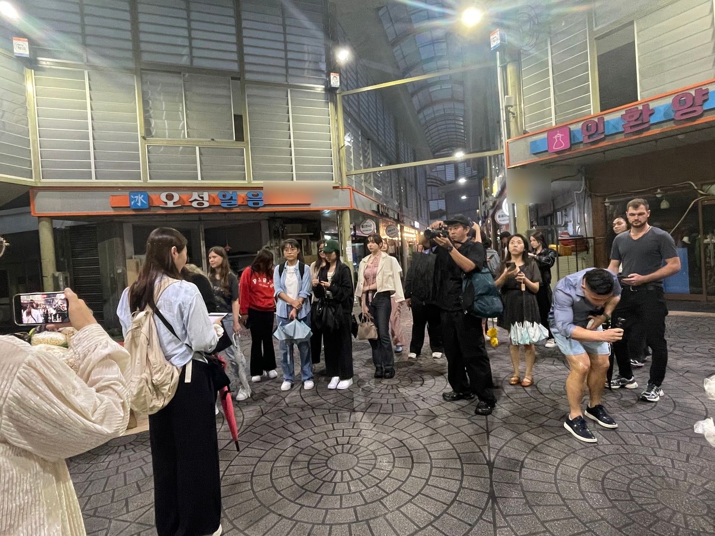 밤 시장 아케이드 골목에 외국인 참가자들이 모여 서 있고 한 사람이 휴대폰으로 촬영하고 있다