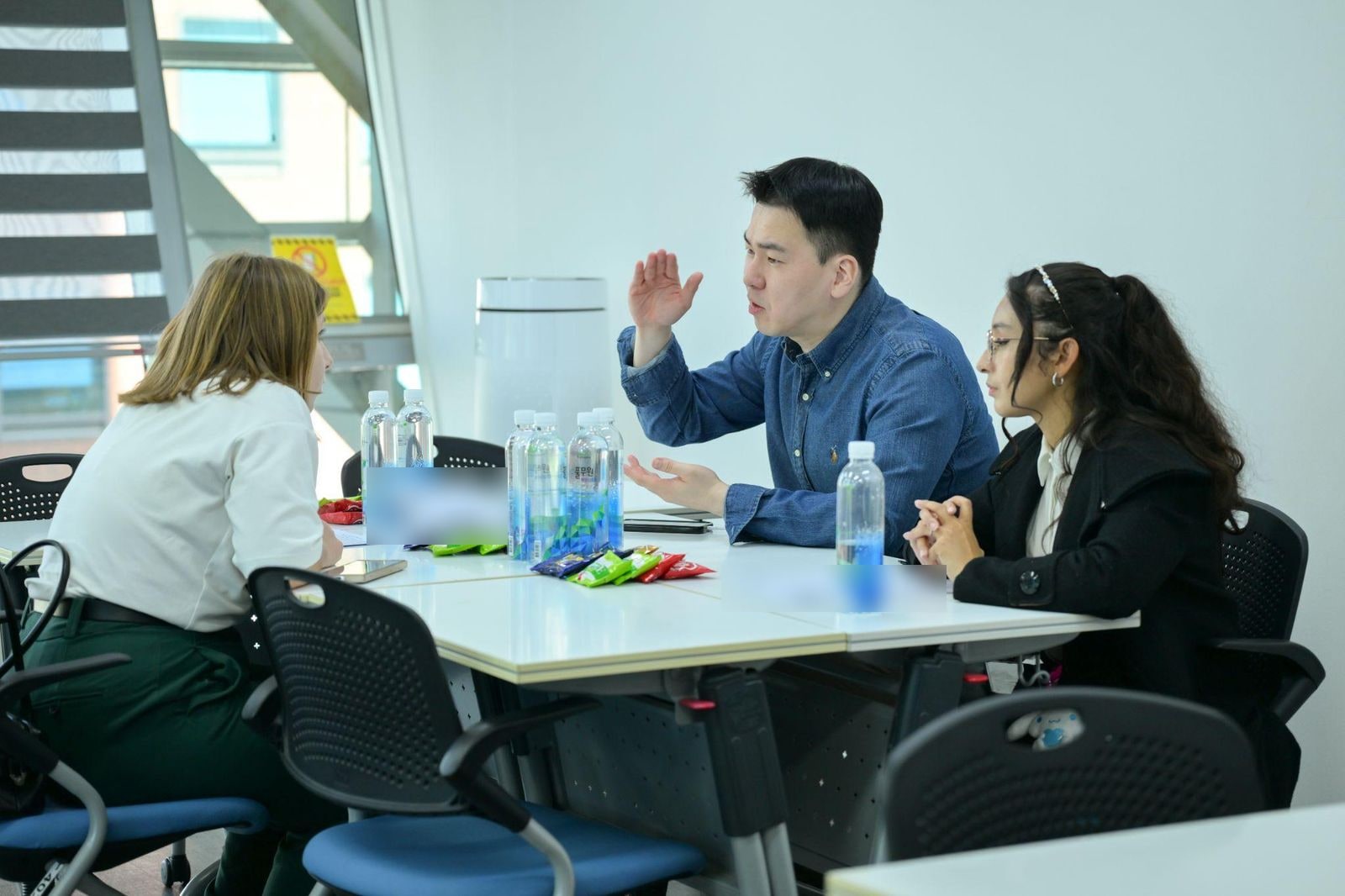 회의실 테이블에 마주 앉은 한국인 담당자가 손짓하며 설명하고, 양옆에 외국인 참가자 두 명이 이야기를 듣고 있다. 테이블 위 서류의 개인정보는 흐리게 처리되어 있다.
