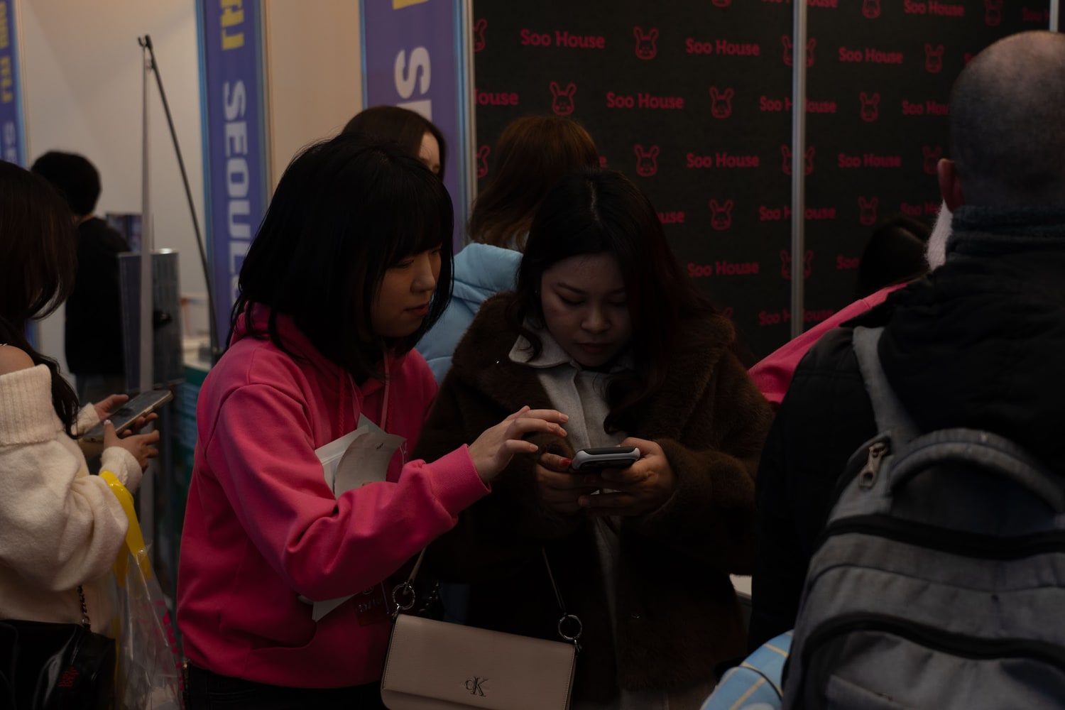행사 부스 앞에서 두 사람이 나란히 서서 한 사람의 휴대폰 화면을 함께 들여다보고 있다. 뒤쪽 벽면에 Soo House 로고가 반복 인쇄돼 있다.
