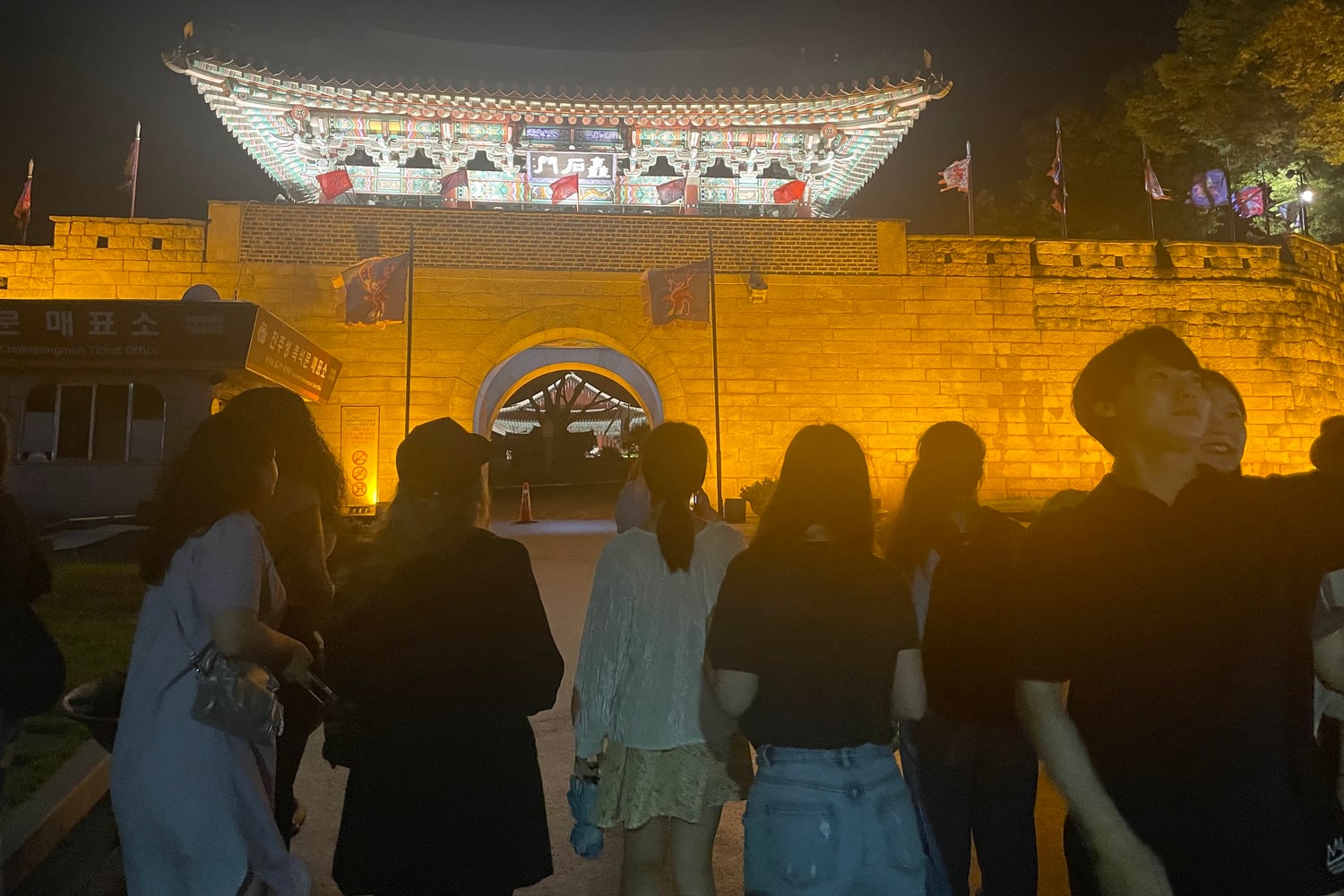 야간 조명이 켜진 성문 앞 광장에 여러 사람이 모여 서 있고, 왼쪽에 매표소 건물이 보인다
