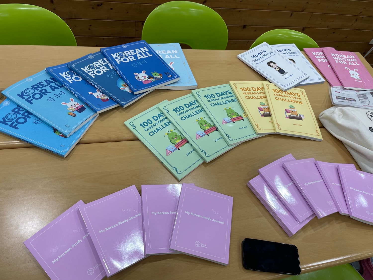 나무 테이블에 한국어 교재가 색깔별로 펼쳐져 있다. 표지에 KOREAN FOR ALL, 100 DAYS KOREAN GRAMMAR CHALLENGE, My Korean Study Journal 이라고 적혀 있다.