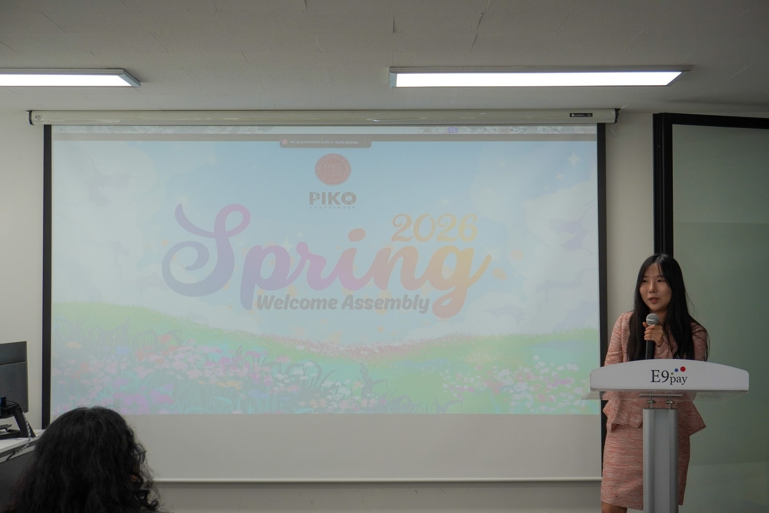 강의실에서 한 진행자가 마이크를 들고 서 있고, 뒤 스크린에 PIKO 2026 Spring Welcome Assembly 라고 적힌 슬라이드가 떠 있다