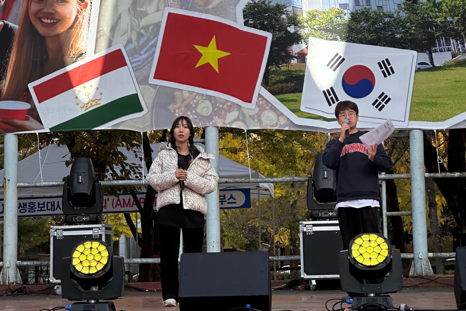 대학 국제학생 축제 무대 위, 배경 현수막에 타지키스탄·베트남·대한민국 국기가 걸려 있고 두 명의 진행자가 마이크를 들고 서 있다