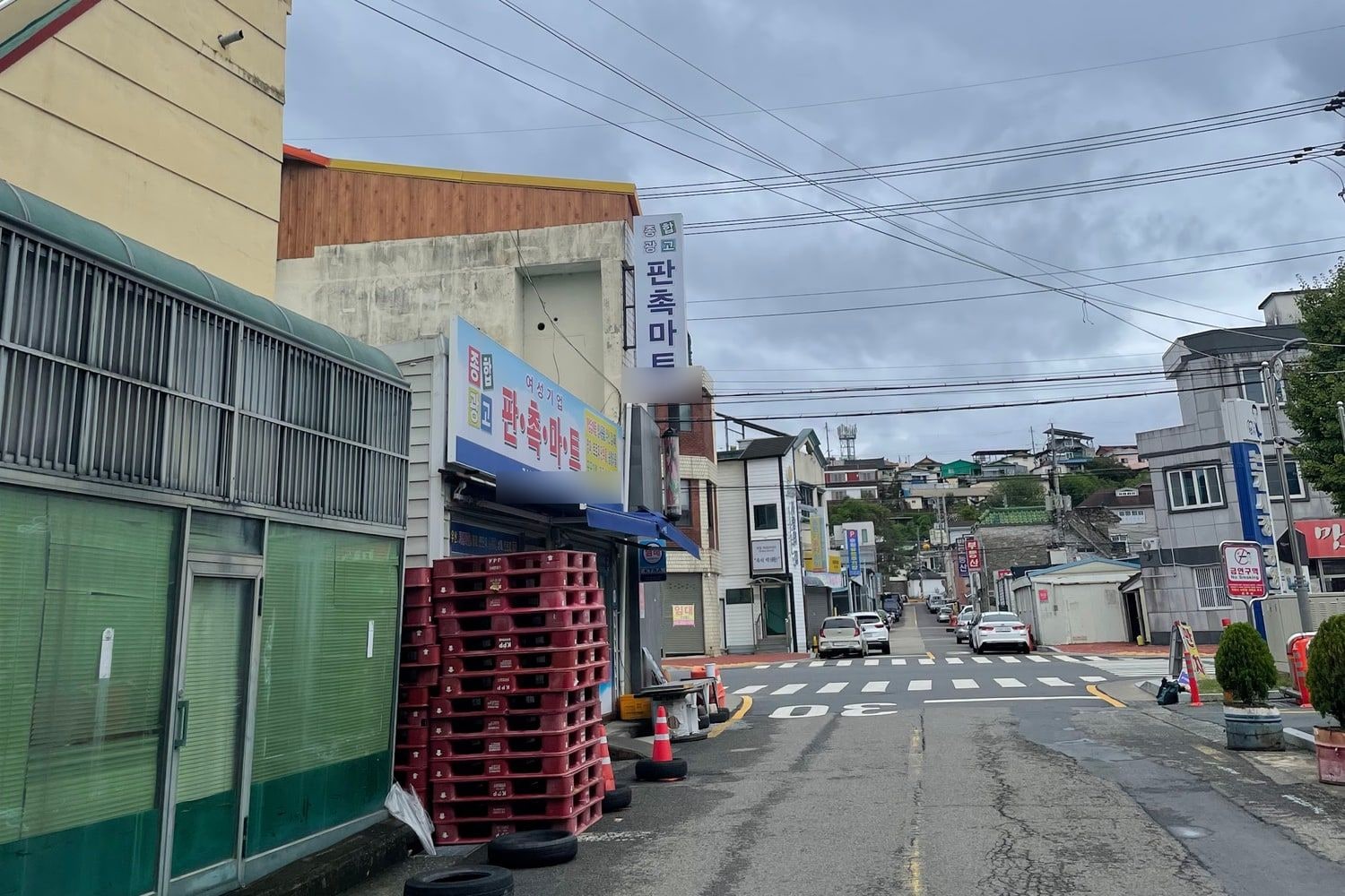경남 진주의 한적한 상가 거리. 왼쪽에 종합광고·판촉물 간판을 단 작은 점포와 쌓아 둔 붉은 플라스틱 상자가 있고, 오른쪽 언덕 위로 주택가가 보인다. 사람은 보이지 않는다.