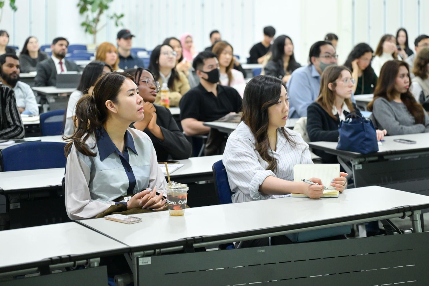 강의실 책상에 앉은 다양한 국적의 참가자들이 앞쪽을 바라보며 설명을 듣고 있고, 일부는 노트에 필기하고 있는 모습.