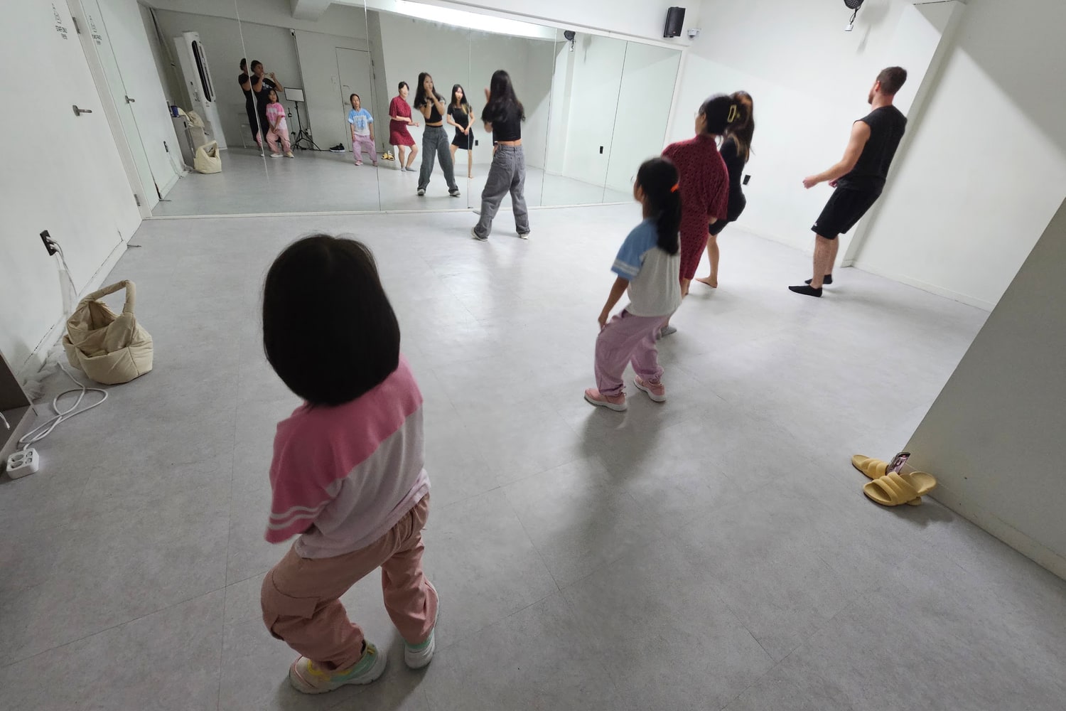 거울이 있는 댄스 스튜디오에서 참가자들이 강사를 향해 서서 동작을 따라 하고 있다. 대부분 카메라를 등지고 있다.