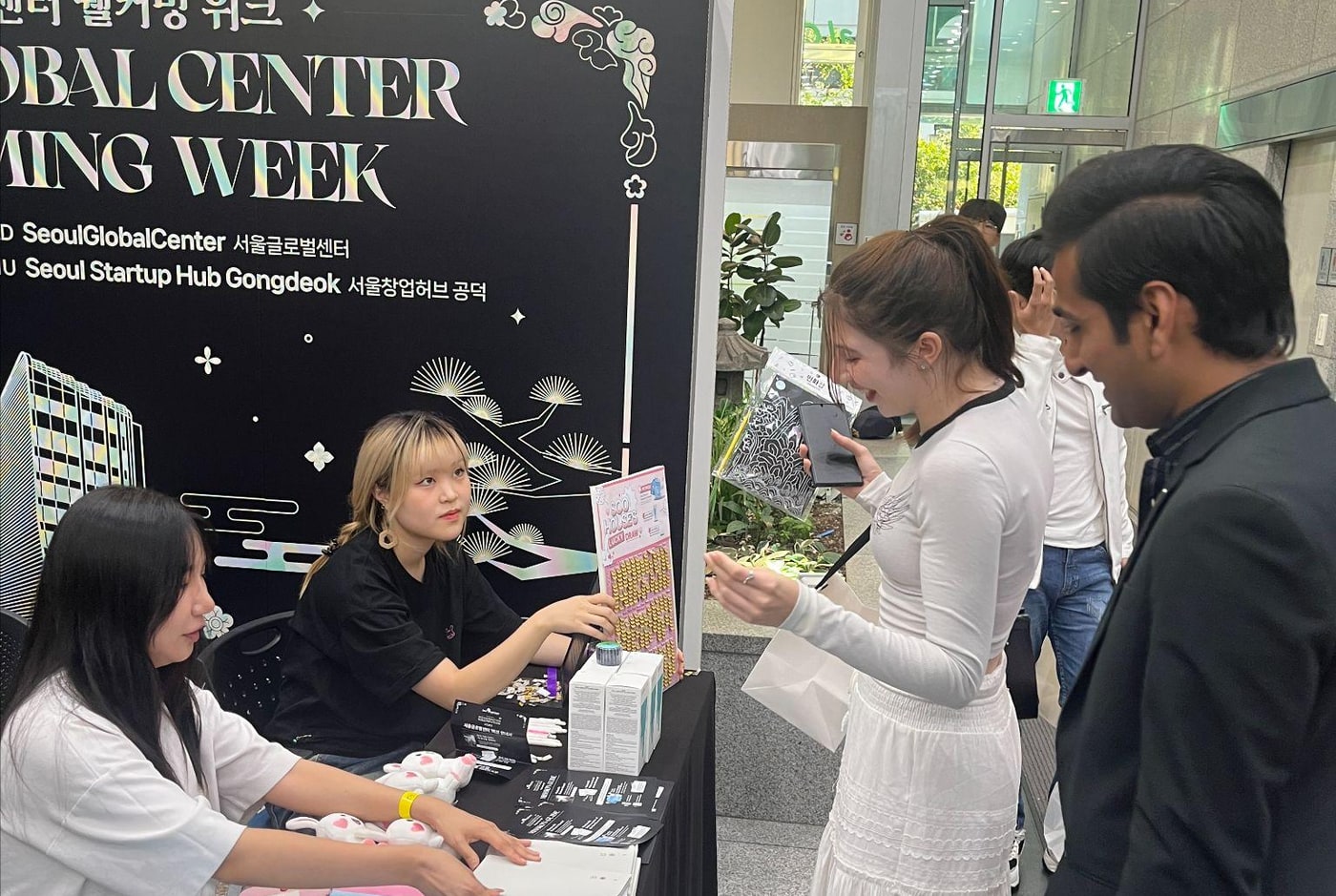 'GLOBAL CENTER WELCOMING WEEK, 서울글로벌센터, 서울창업허브 공덕'이라고 적힌 검은 배너 앞 부스에서, 스태프가 외국인 참가자에게 분홍색 수하우스 안내물을 건네고 있는 모습.