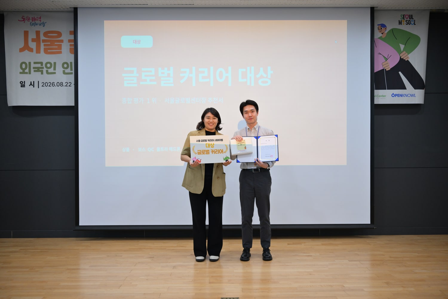 무대에서 두 사람이 글로벌 커리어 대상 보드와 추천서를 들고 서 있고, 뒤 화면에 글로벌 커리어 대상이라는 문구가 떠 있는 모습