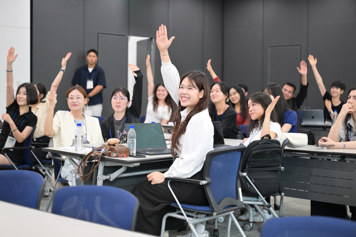 강의실에서 여러 참가자가 동시에 손을 들고 있고 가운데 참가자가 웃고 있는 모습