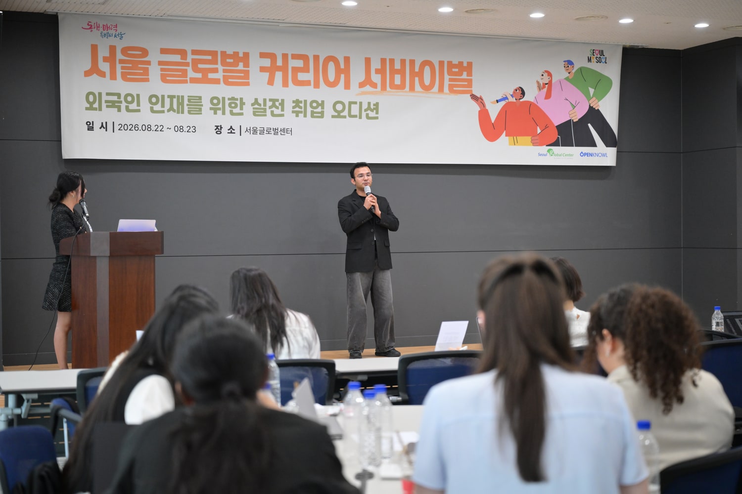 행사 배너가 걸린 무대에서 참가자가 마이크를 들고 이야기하고, 객석의 참가자들이 이를 지켜보는 모습