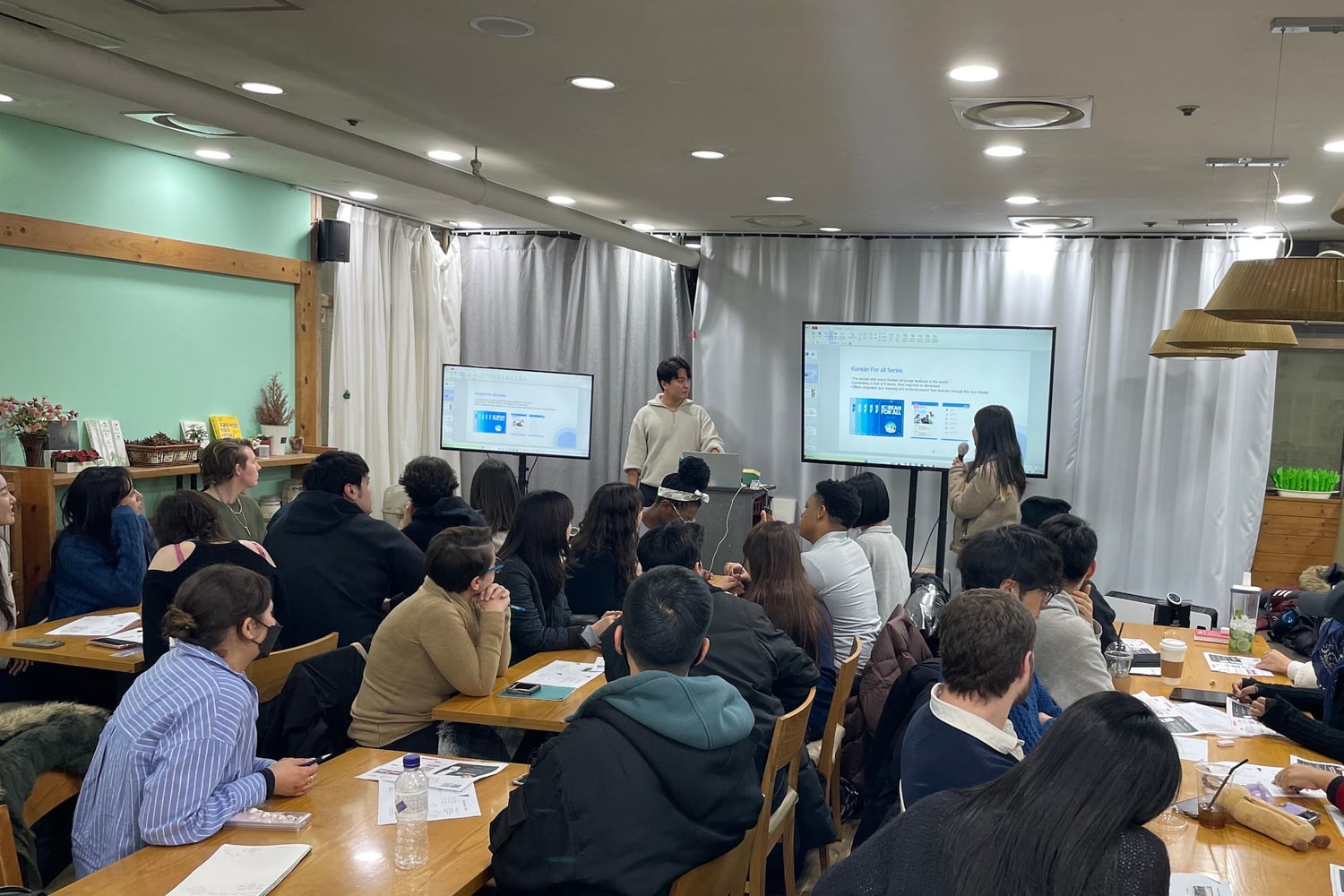 한국어 클래스 현장. 진행자 두 사람이 앞에 서 있고 참가자들이 긴 테이블에 앉아 화면을 보고 있다. 화면에는 Korean For all Series 라고 적혀 있다.