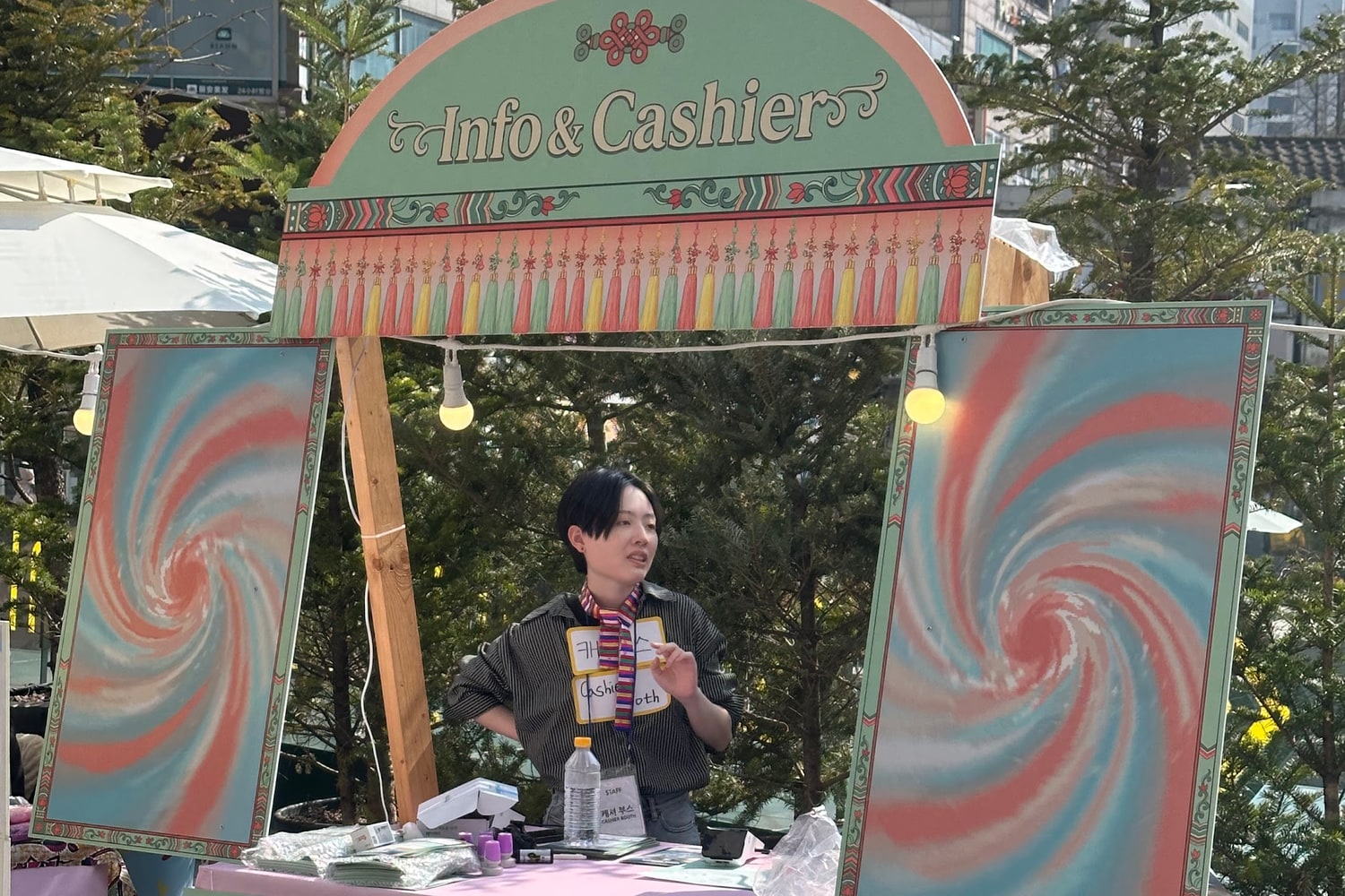 야외 행사장에 세워진 Info & Cashier 안내 부스. 한국 전통 문양으로 장식한 아치형 간판 아래에서 스태프 한 명이 방문객 쪽을 보며 설명하고 있고, 목에 건 이름표에는 개인 이름이 아니라 캐셔 부스라는 역할이 적혀 있다.