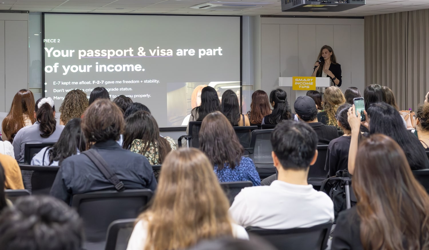 수하우스 세션 현장. 스크린에 “Your passport & visa are part of your income.”이라는 문장과 “E-7 kept me afloat. F-2-7 gave me freedom + stability.”라는 문구가 떠 있고, 객석은 참가자들로 채워져 있다.