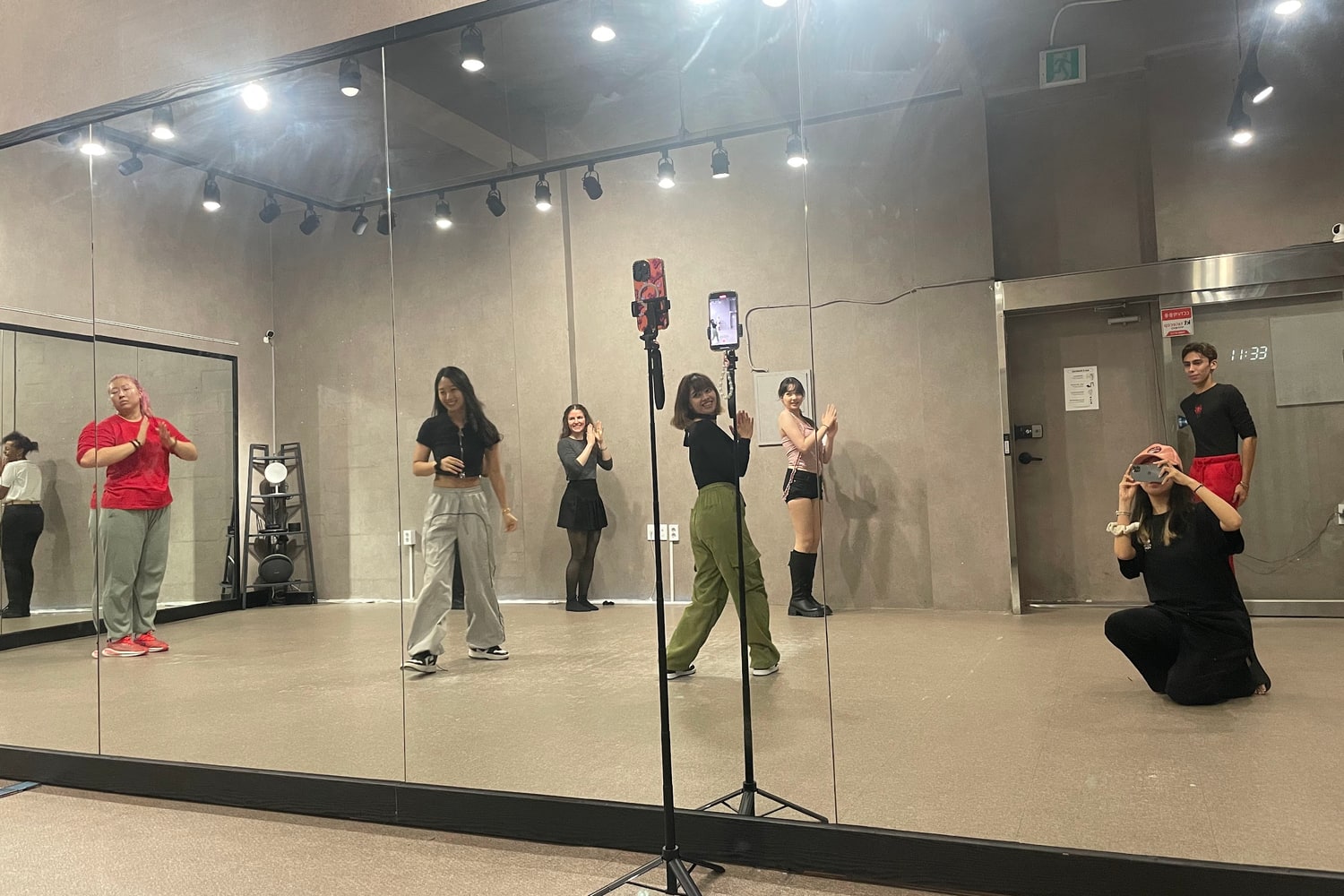 거울이 있는 댄스 스튜디오에서 여러 국적의 참여자들이 강사를 따라 동작을 맞추고 있고 오른쪽 벽 시계는 11시 33분을 가리키고 있다