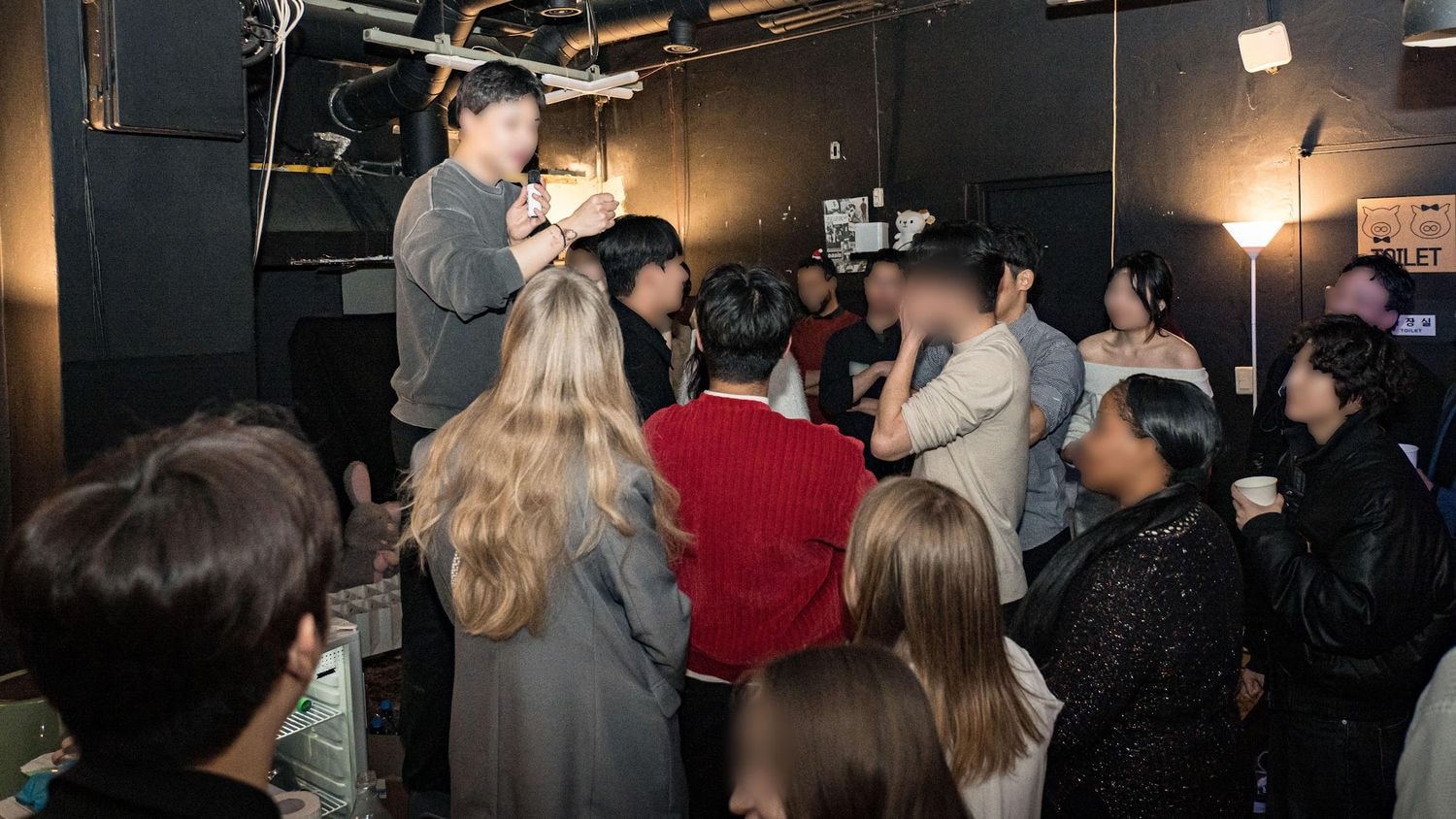 어두운 지하 공간에서 마이크를 든 진행자를 참가자 여러 명이 둘러서서 바라보고 있다. 대부분 뒷모습이고 얼굴은 흐리게 처리돼 있다.